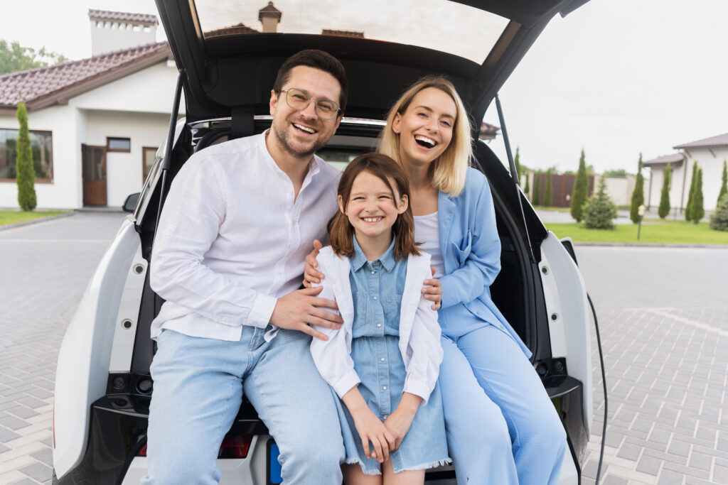 Happy Family Sitting in Car Trunk Outside Modern House
